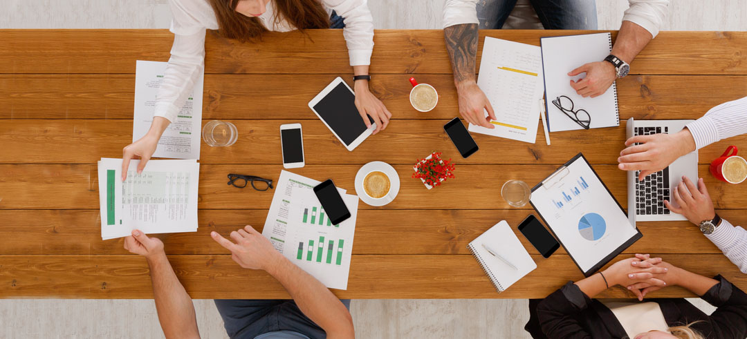 Mesa vista desde arriba de varias personas reunidas con móviles, tablet y ordenador